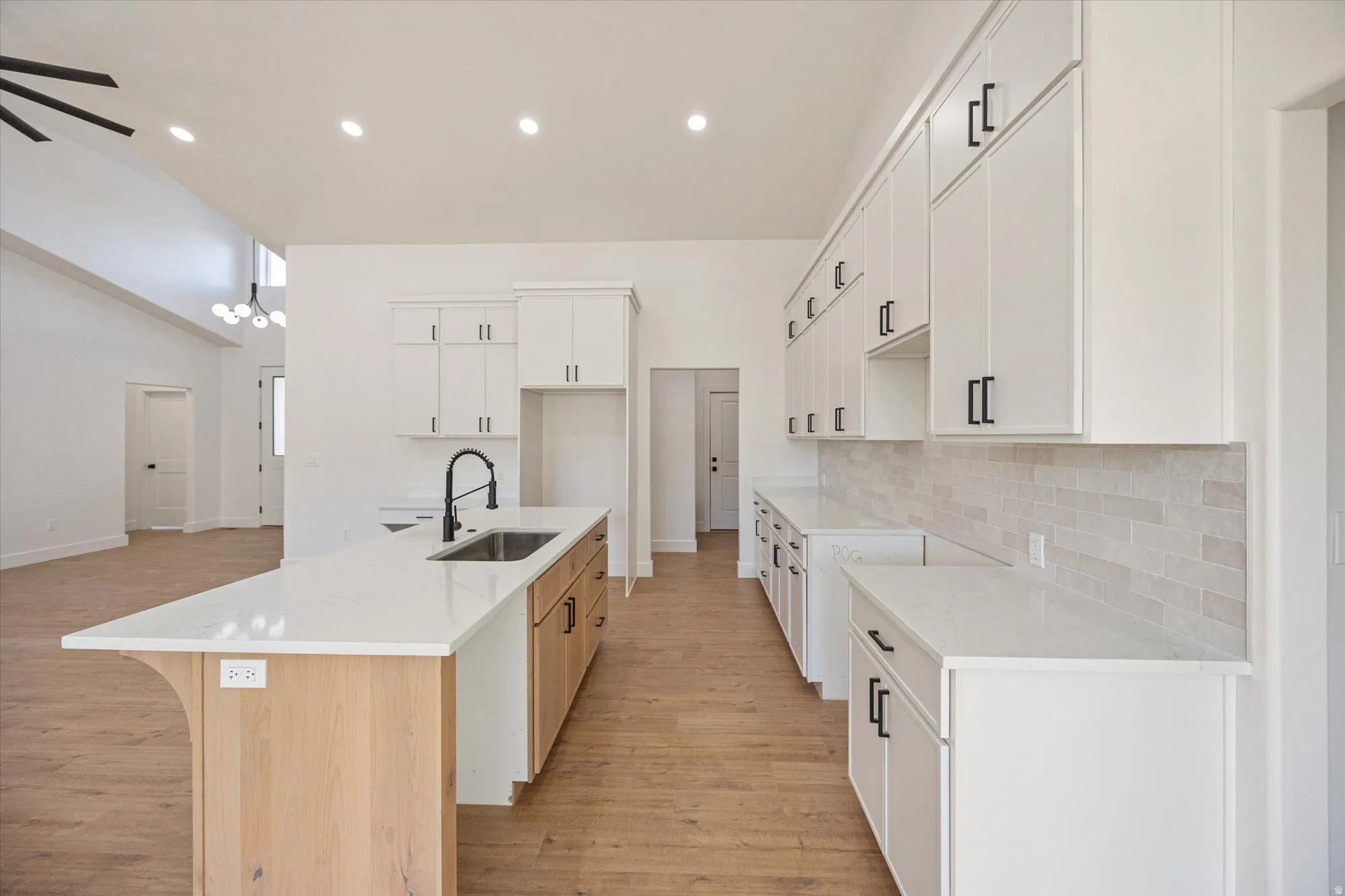 Spacious kitchen featuring an extended island with a built-in sink and seating overhang, white shaker-style cabinetry with matte black hardware, light-toned wood-finish flooring, white countertops, and a full-height tile backsplash