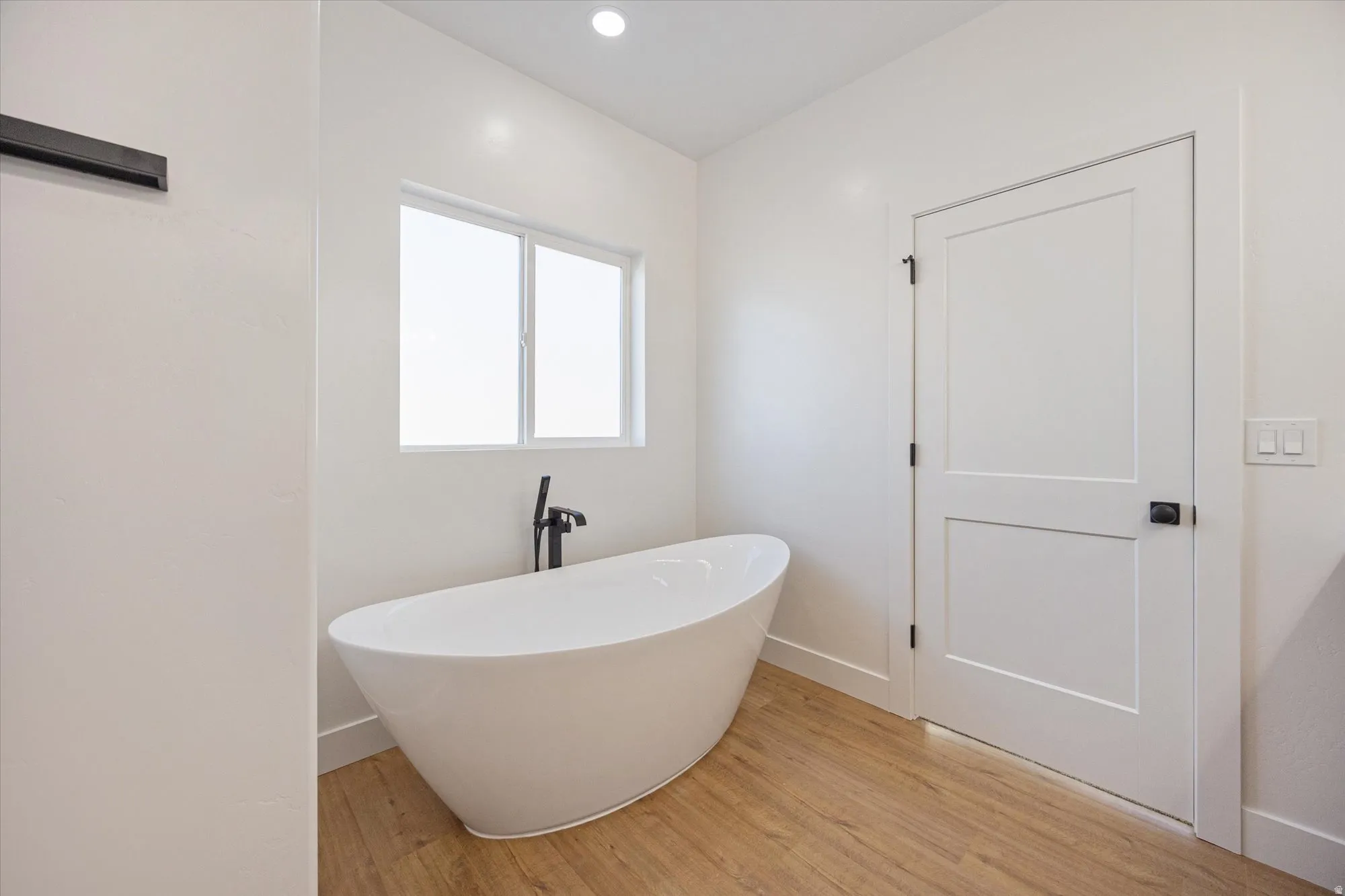 Freestanding soaking tub with a matte black floor-mounted faucet, wood-finish flooring, recessed lighting, and a modern two-panel interior door with matte black hardware