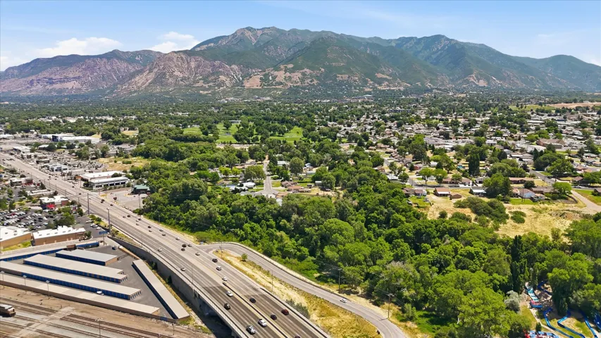 Expansive aerial perspective showcasing a mountainous backdrop, a multi-lane highway, a residential area with mature trees, commercial buildings, and a large facility with multiple storage units