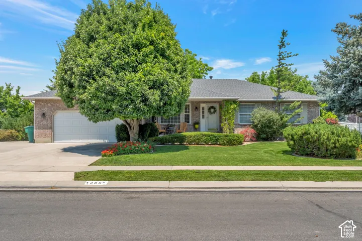 Brick-clad residence featuring a front lawn, mature trees, an attached garage, a paved driveway, and a covered entry