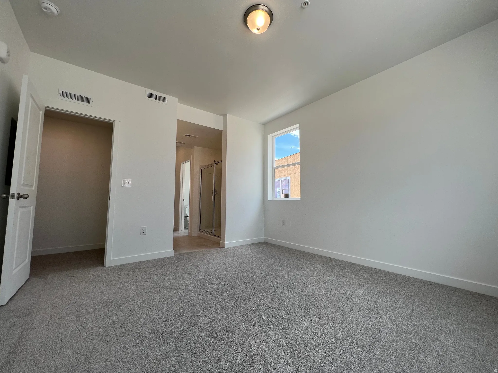 Carpeted room featuring a single window, overhead lighting, and an adjacent open doorway revealing a shower enclosure