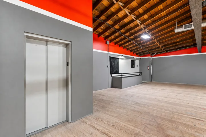 Spacious open-concept interior featuring light wood-finish flooring, exposed wood beam ceilings, and a neutral gray and vibrant red wall palette