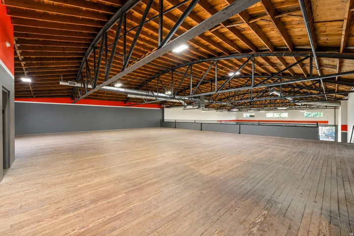 Spacious open area featuring wood-finish flooring, exposed beam ceiling structure, and a mezzanine level with a metal railing