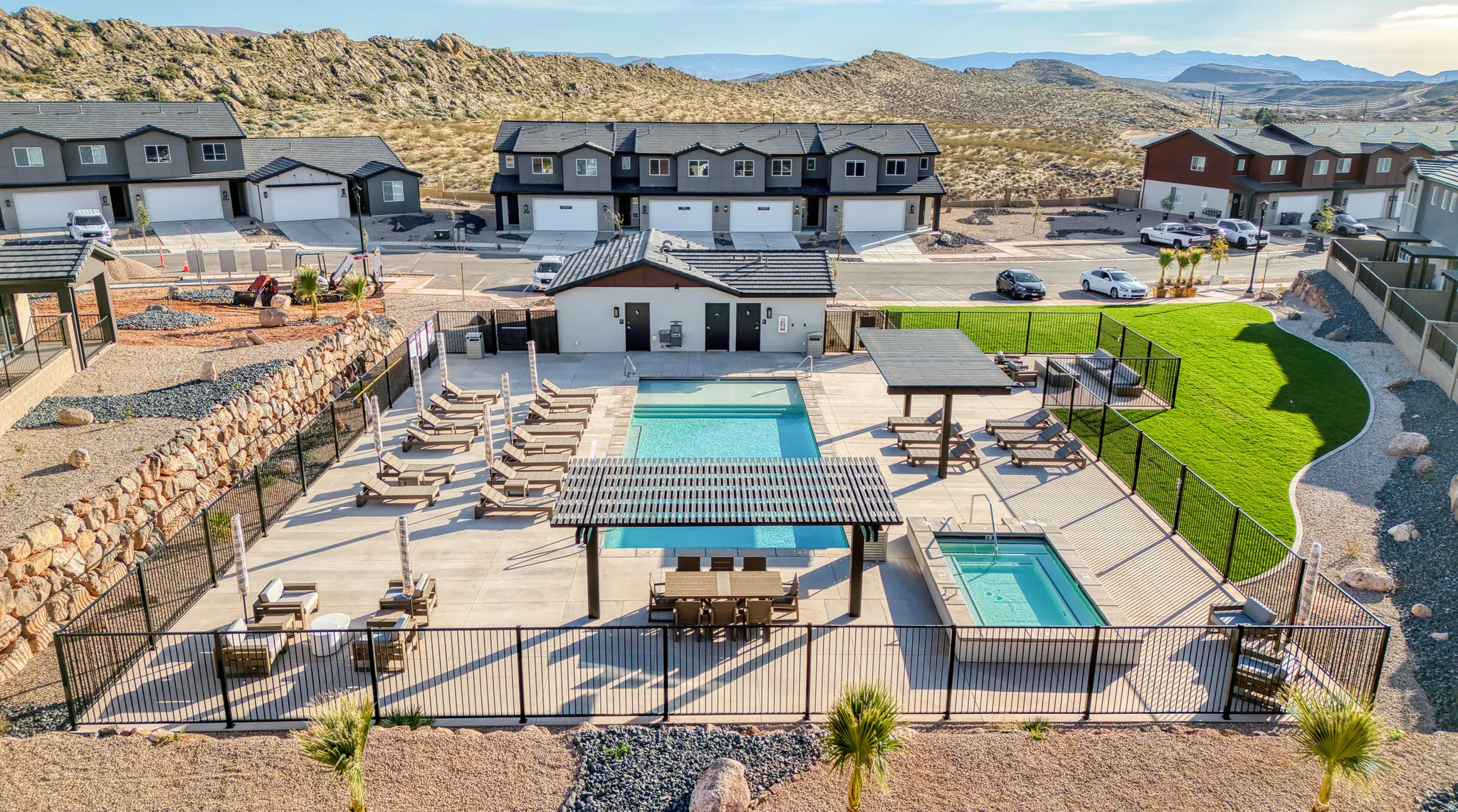 Resort-style outdoor living space featuring a resort-style pool and spa with black safety fencing, expansive sun deck with lounge seating areas, manicured lawn, pergola shade structures, and panoramic desert landscape backdrop with mountain views