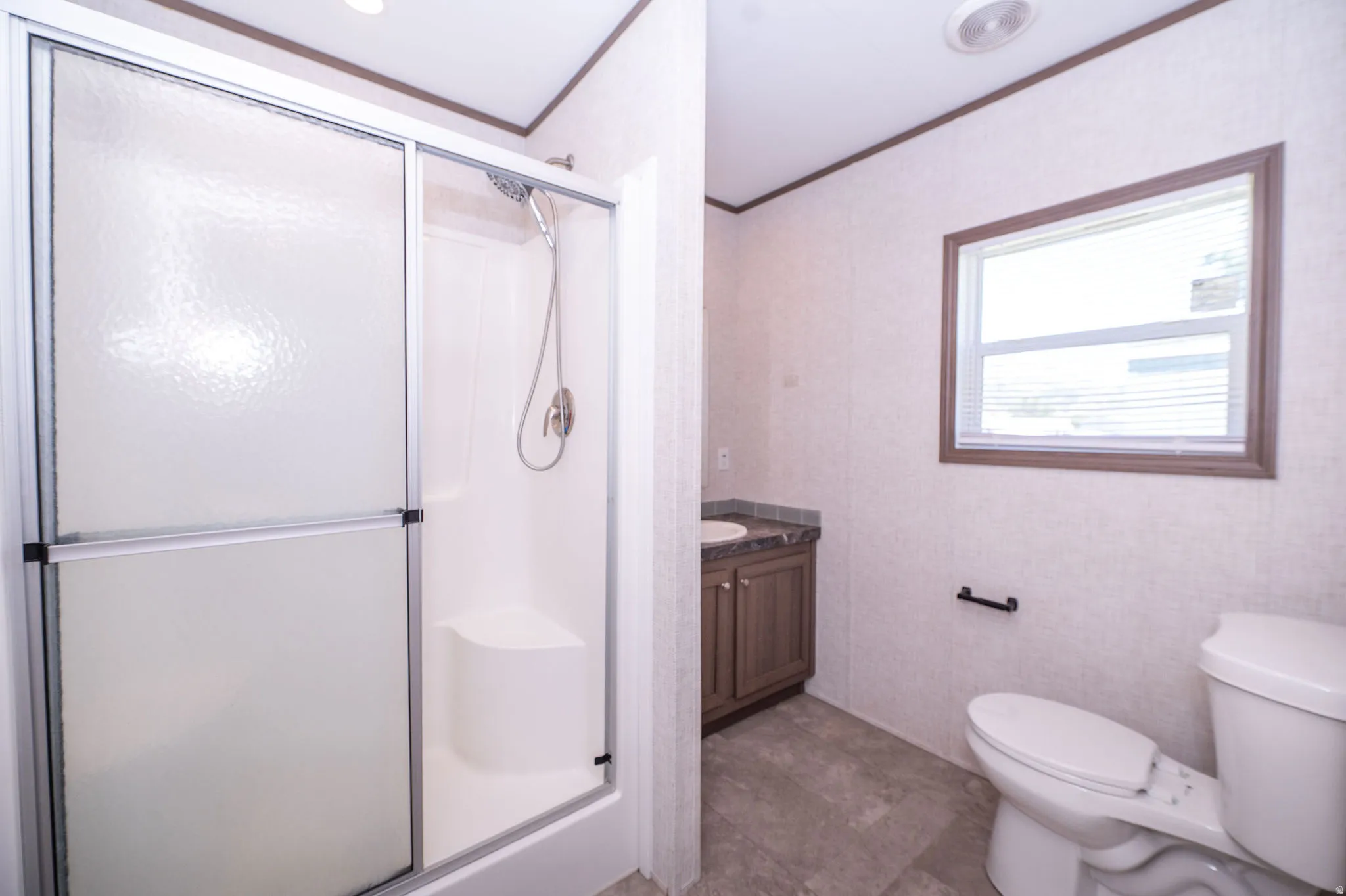 Bathroom featuring a shower with sliding opaque glass doors and a built-in seat, a wood-finish vanity with an integrated sink and dark countertop, a window with horizontal blinds, and a toilet