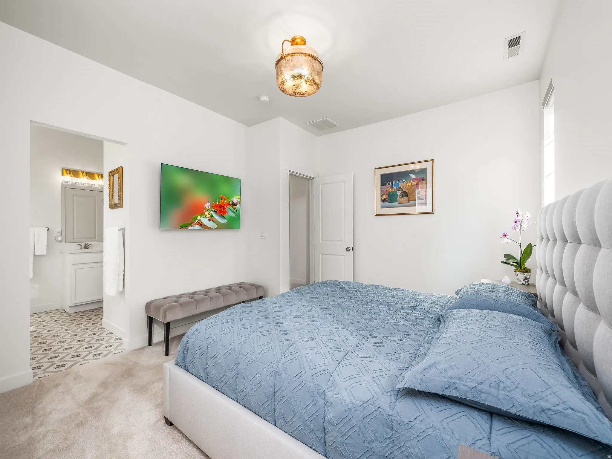 Bedroom with a light gray tufted headboard, soft neutral carpet, and an en-suite bathroom featuring patterned tile flooring and a single vanity with a white countertop