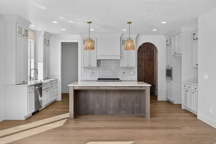 Expansive kitchen featuring a large central island with a light-toned countertop, white shaker cabinetry with gold-toned hardware, stainless steel appliances, and wood-finish flooring