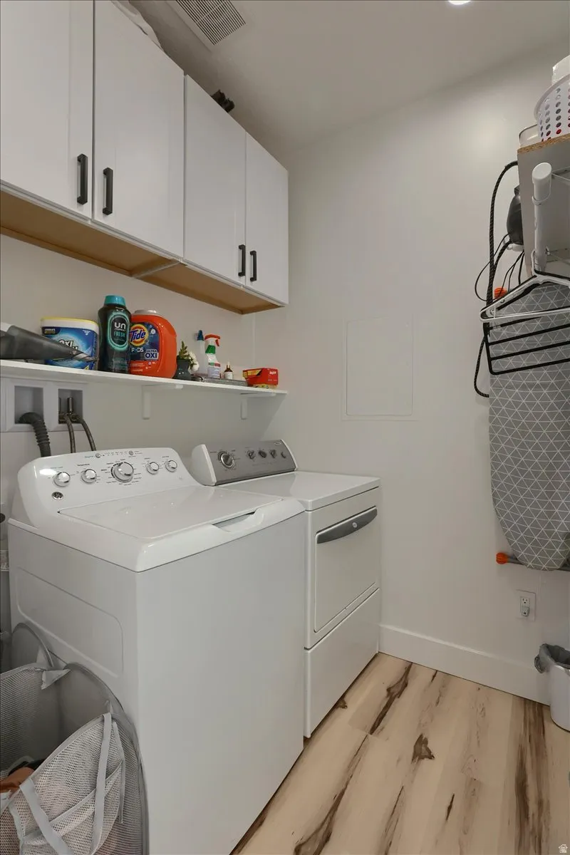 Dedicated laundry area featuring white appliances, upper white cabinetry with dark hardware, a wall-mounted shelving unit, and wood-finish flooring