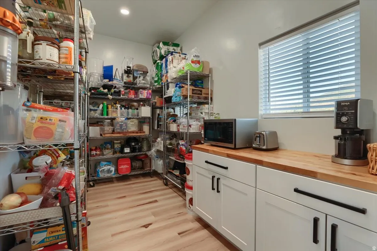 Spacious pantry featuring extensive wire shelving, natural wood-finish countertops, white cabinetry with matte black hardware, wood-finish flooring, and a window with horizontal blinds