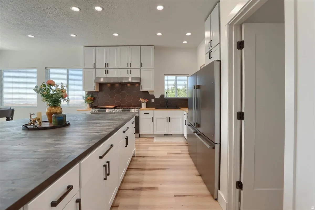 Spacious kitchen featuring an expansive center island with ample storage, light wood-finish flooring, white cabinetry with dark hardware, a hexagonal tile backsplash, and recessed lighting throughout
