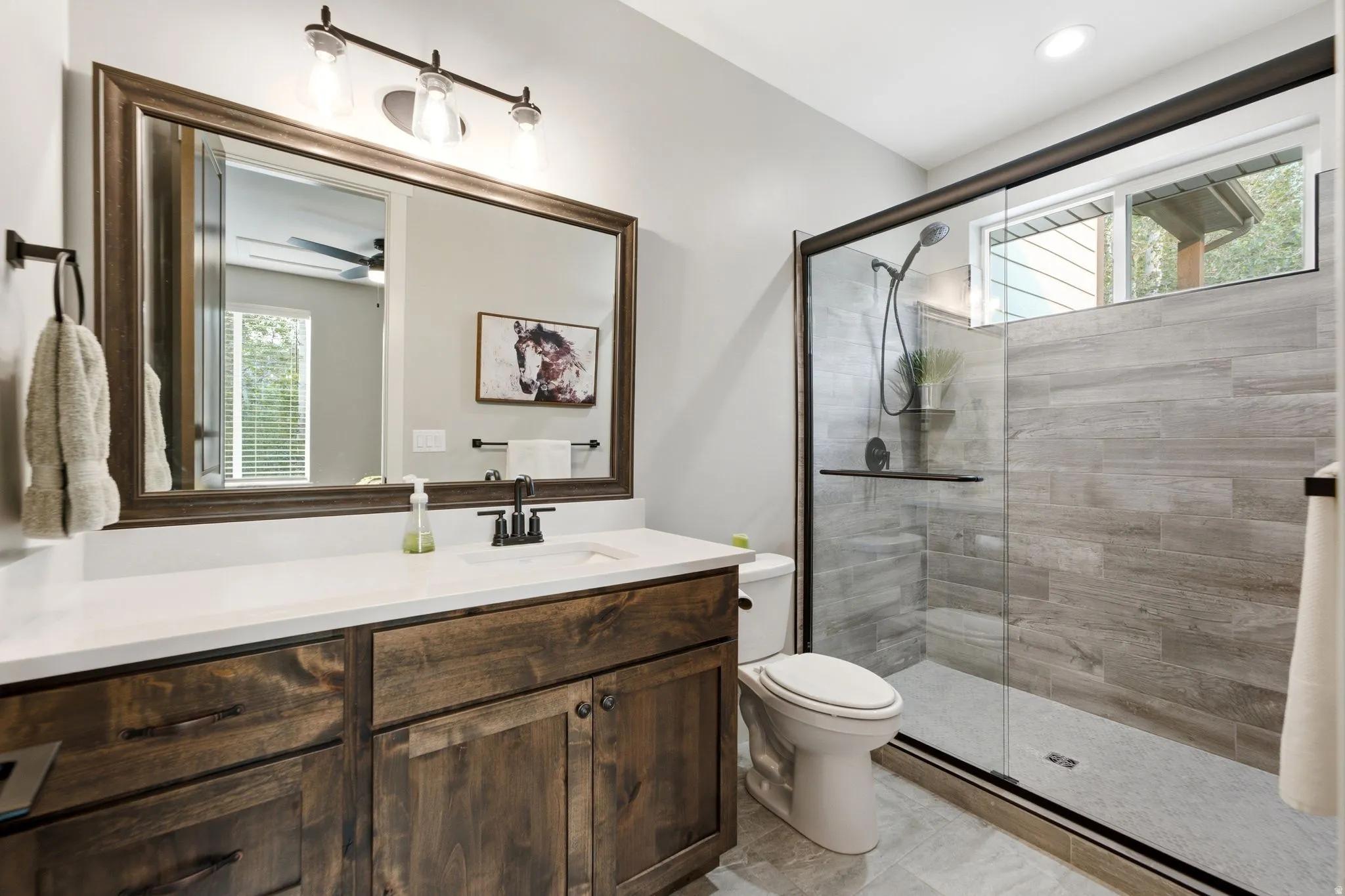 Bathroom featuring a dark wood vanity with a white countertop and undermount sink, framed mirror with three-light fixture, and a glass-enclosed shower with gray tiled walls and a transom window