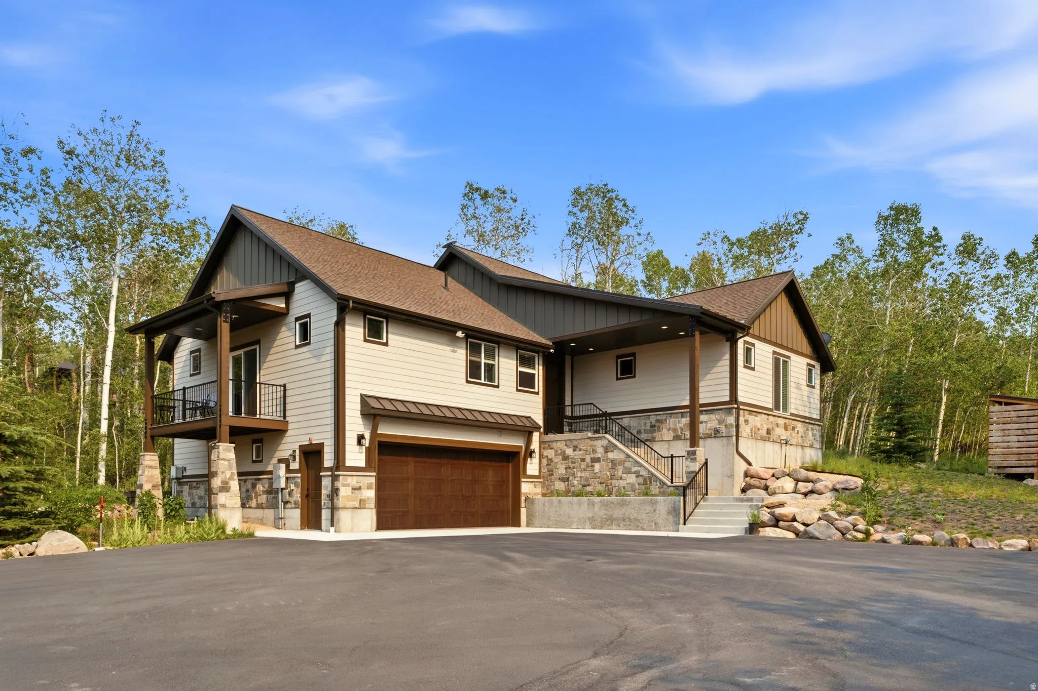 Modern mountain residence featuring a gabled roofline, stone and siding exterior, an attached garage with a wood-finish door, a covered porch entry, and an upper-level balcony with black railing