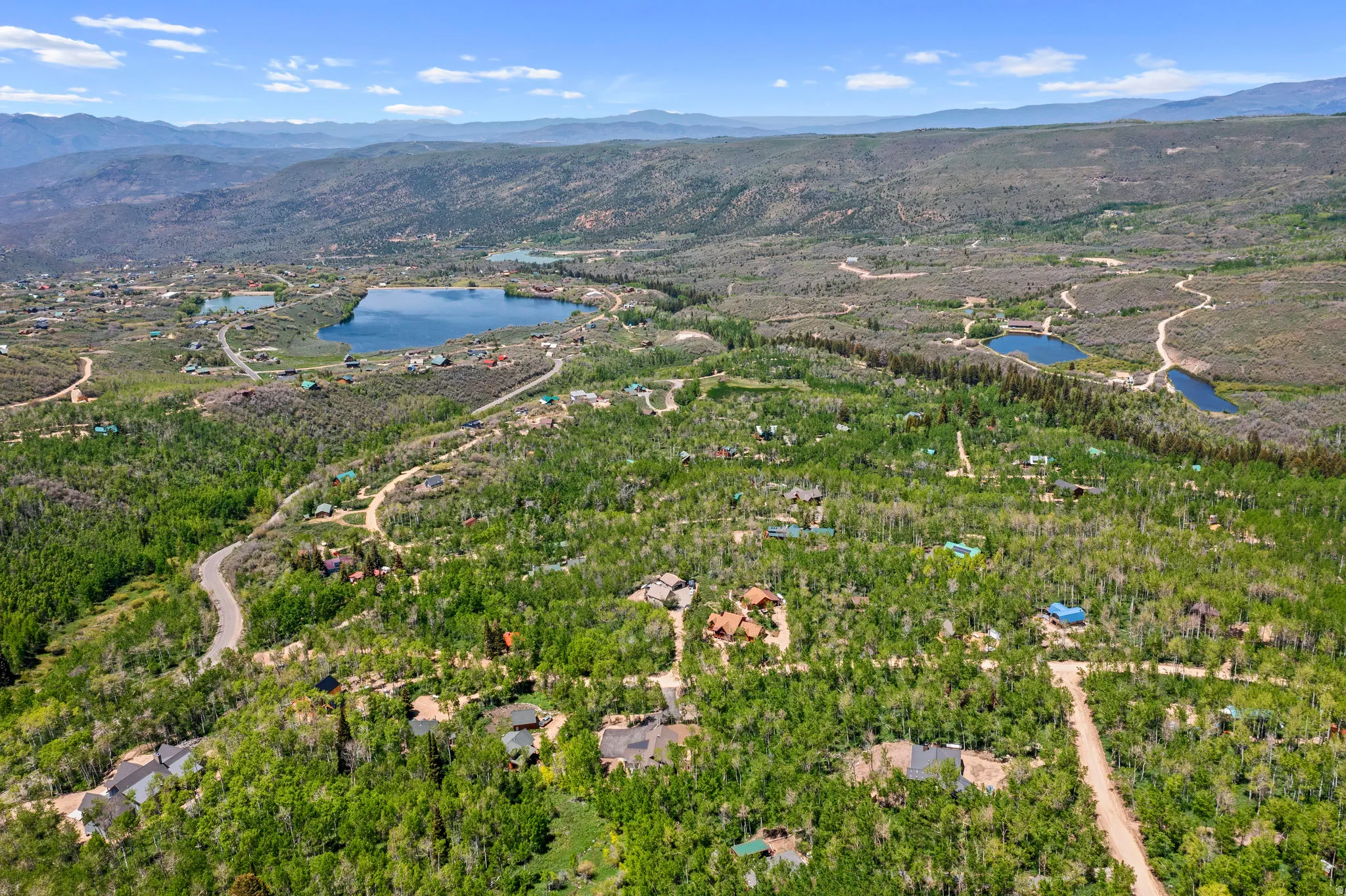 Expansive aerial landscape featuring a large lake, surrounding forested terrain, winding roads, and distant mountain ranges under a clear sky