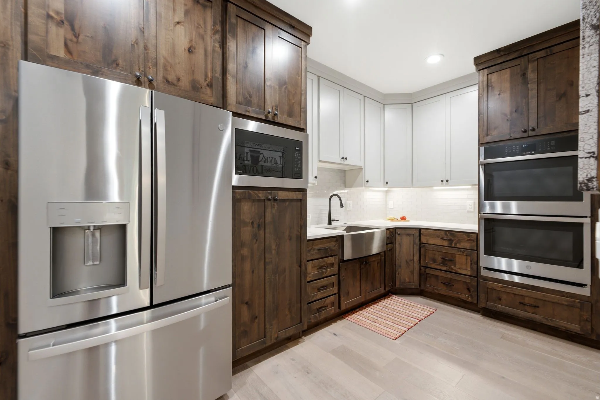 Kitchen featuring stainless steel appliances, a farmhouse sink, white subway tile backsplash, and two-tone cabinetry with dark wood-finish lower cabinets and light-toned upper cabinets