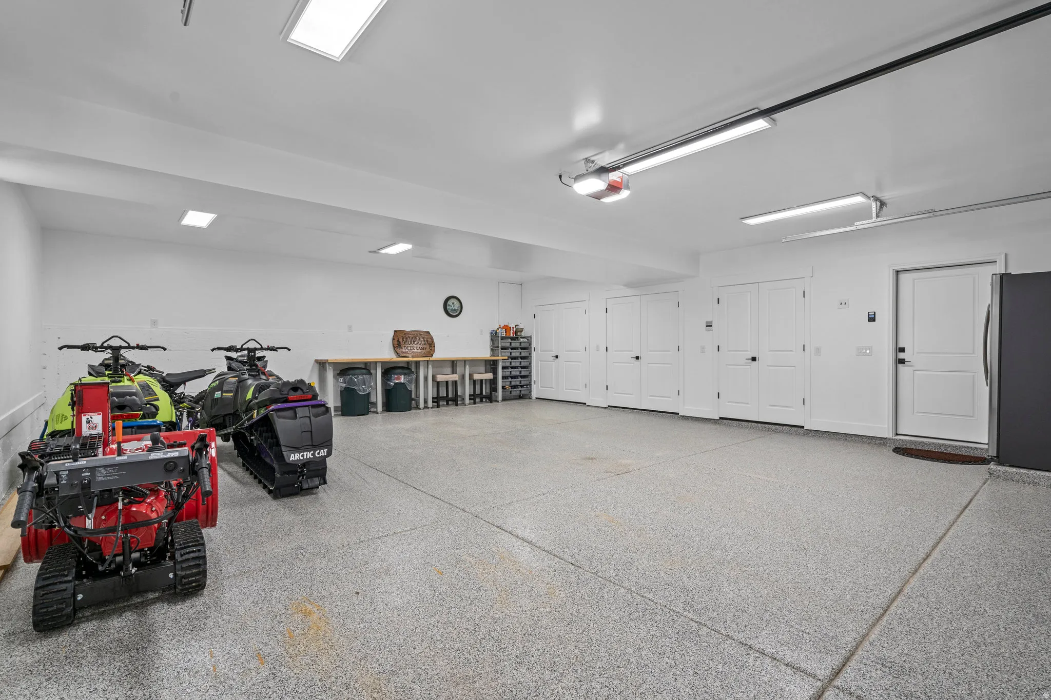 Spacious garage featuring an epoxy-coated concrete floor, bright overhead lighting, multiple built-in storage closets with white panel doors, a workbench area, and an additional access door