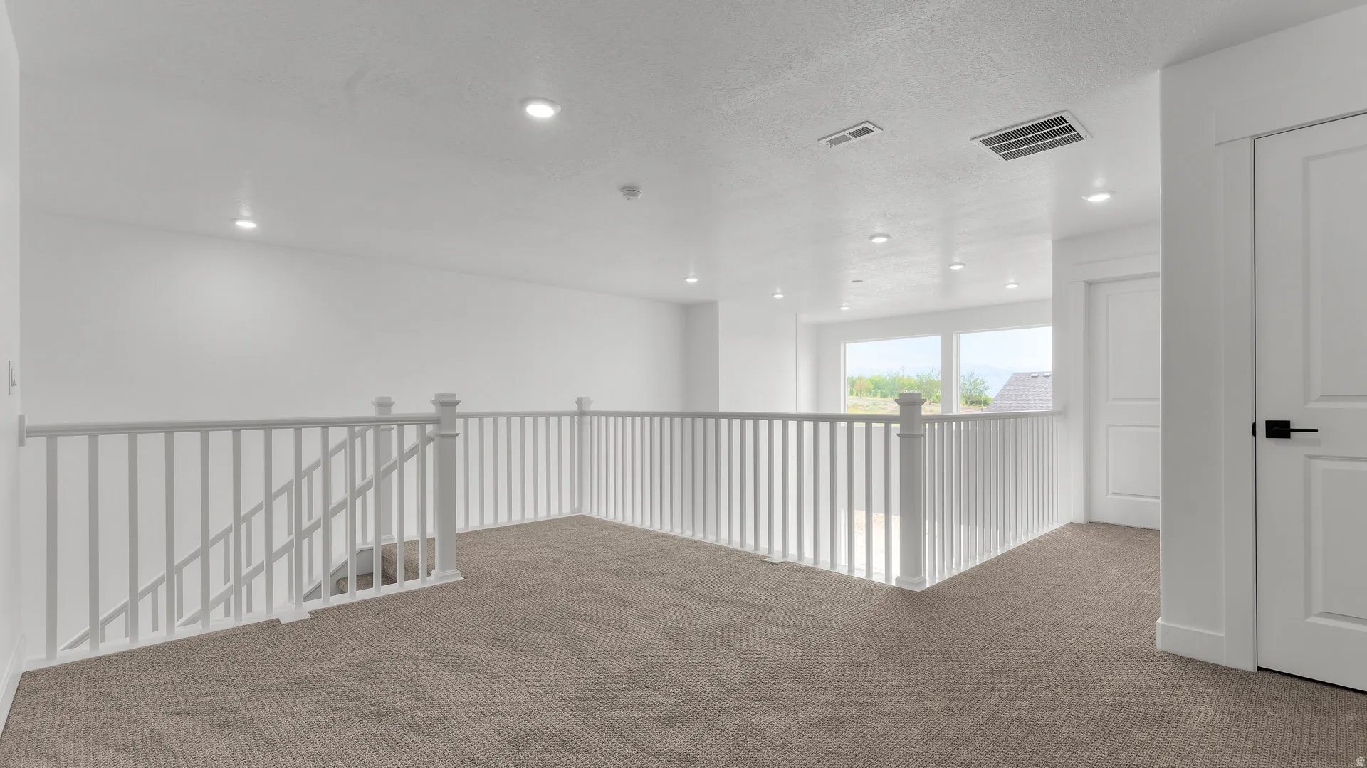 Expansive carpeted loft area featuring white railings, recessed lighting, and large windows providing abundant natural illumination