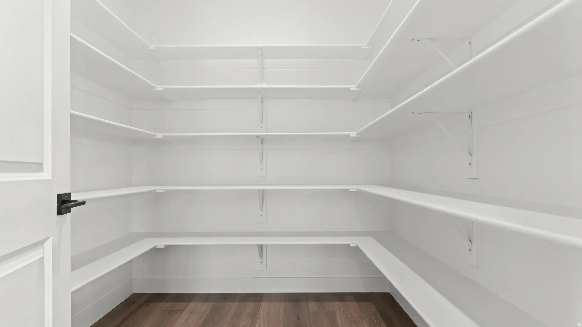 Walk-in pantry with extensive white shelving, wood-finish flooring, and a modern black door handle