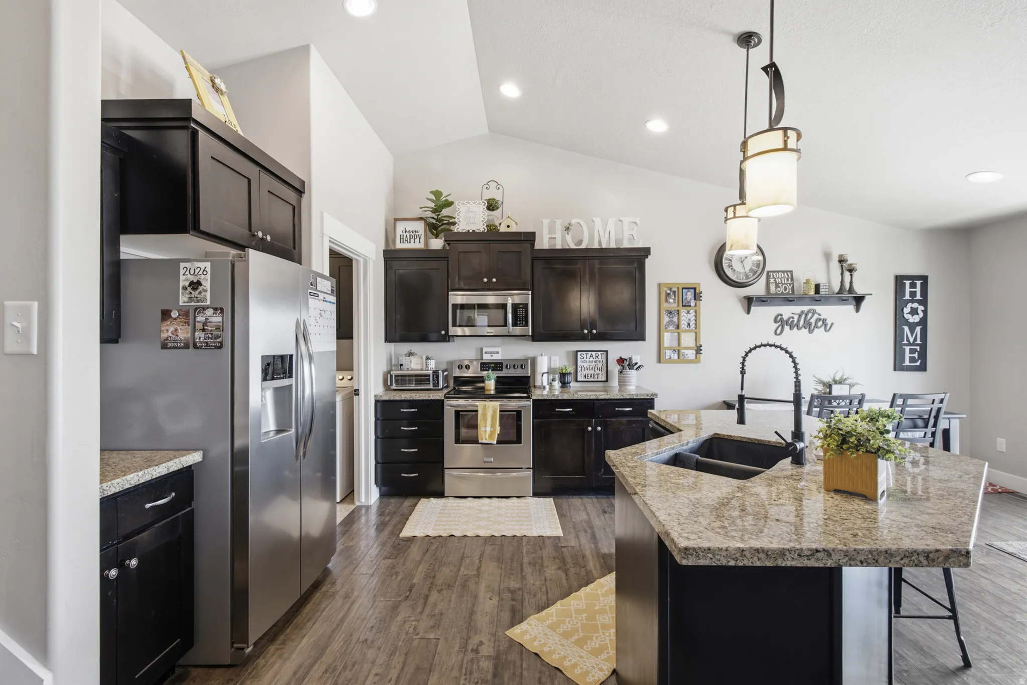 Kitchen featuring wood-finish flooring, dark cabinetry, stainless steel appliances, and a central island with a double basin sink and stone-finish countertop