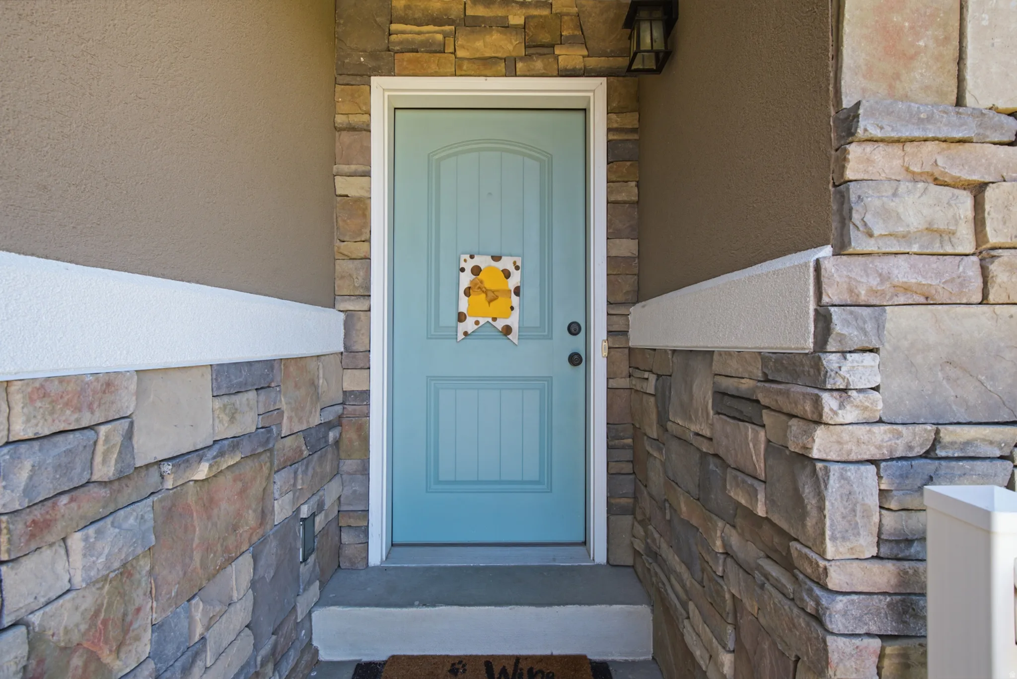 Inviting entryway featuring a paneled turquoise door, stacked stone accents, stucco siding, and an exterior wall lantern
