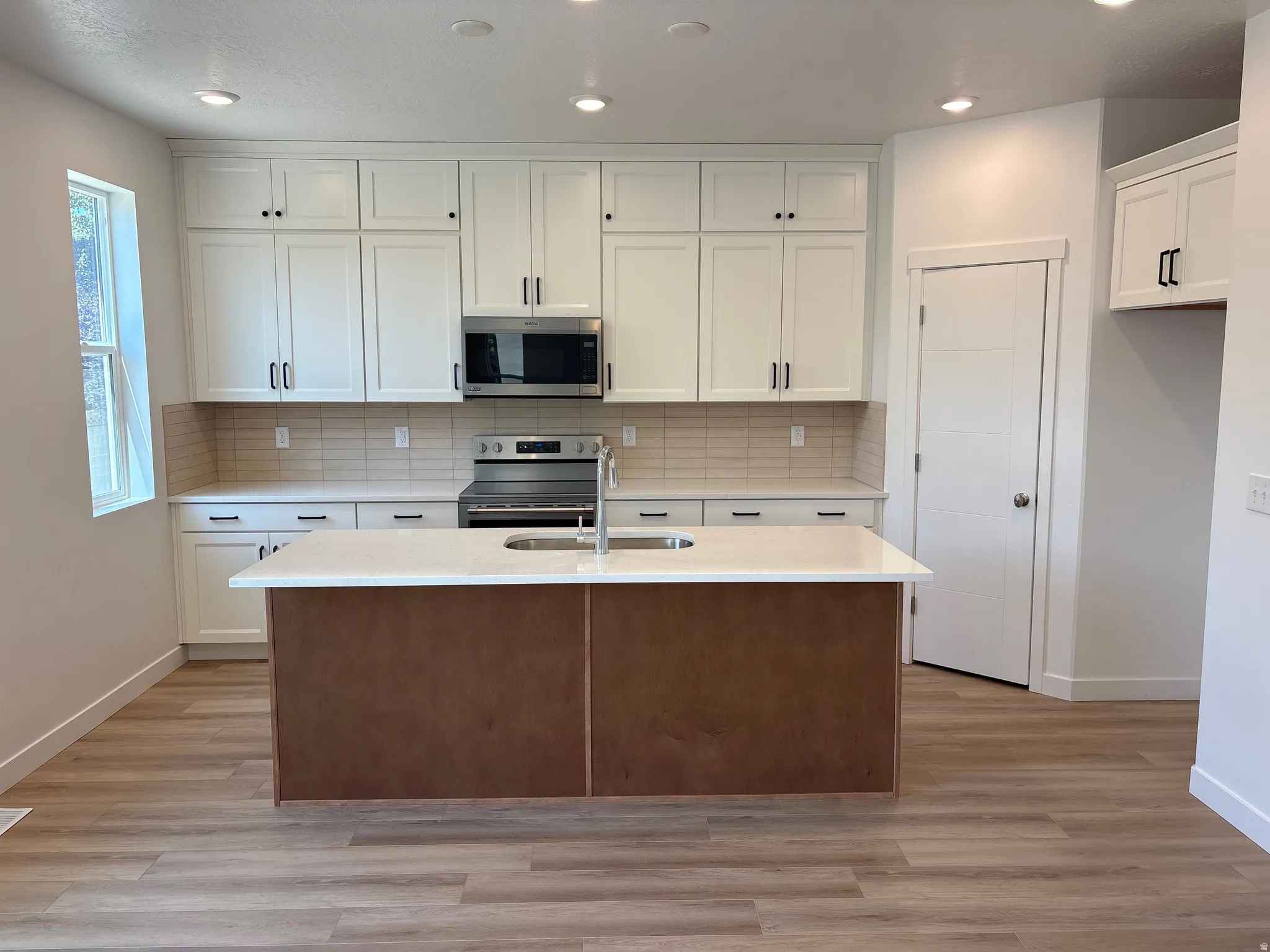 Contemporary kitchen featuring a large center island with a sink, white cabinetry, integrated stainless steel appliances, light-toned backsplash tiling, and wood-finish flooring