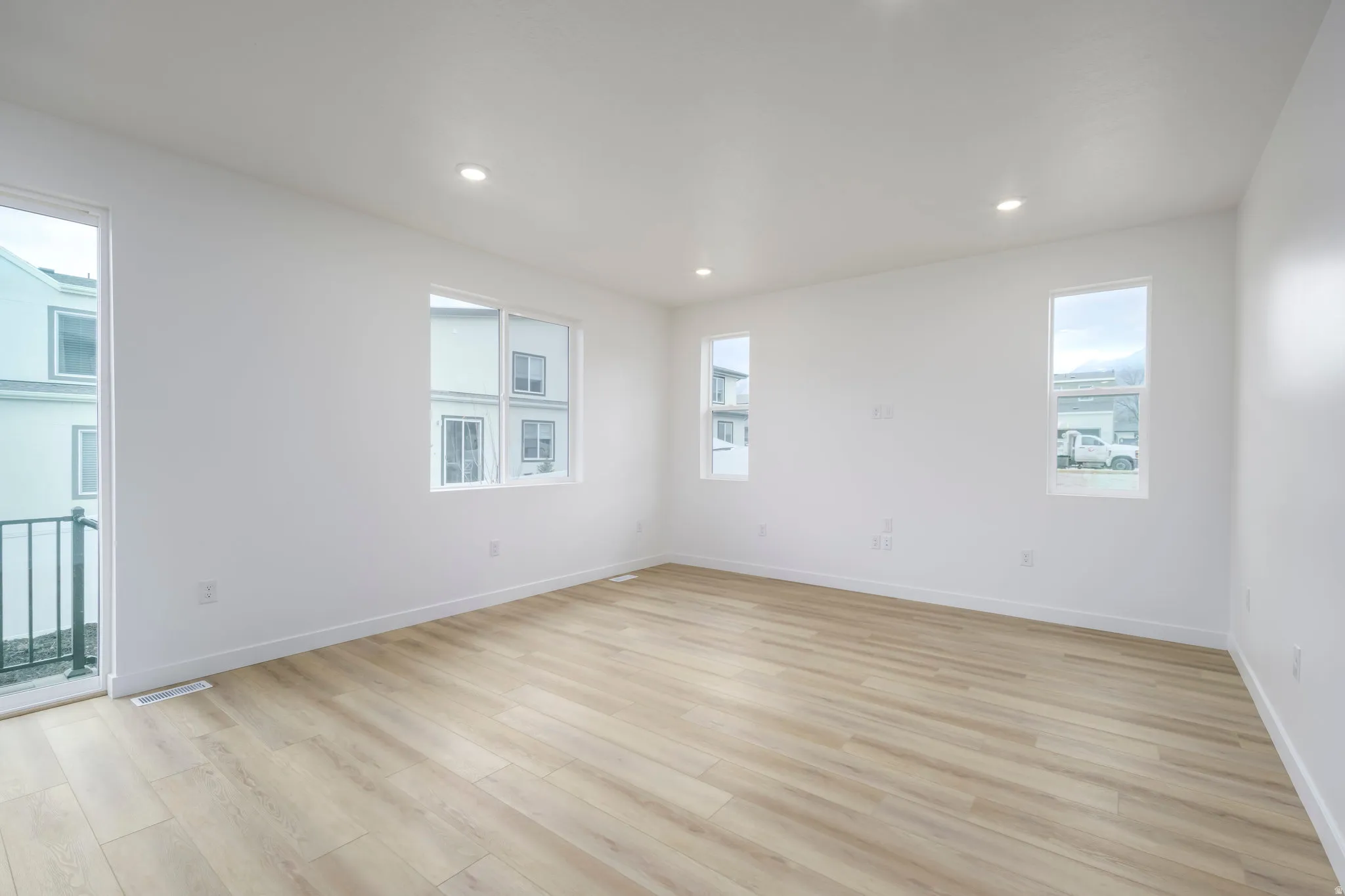 Spacious room featuring light wood-finish flooring, recessed lighting, multiple windows providing natural illumination, a sliding glass door with balcony access, and crisp white walls