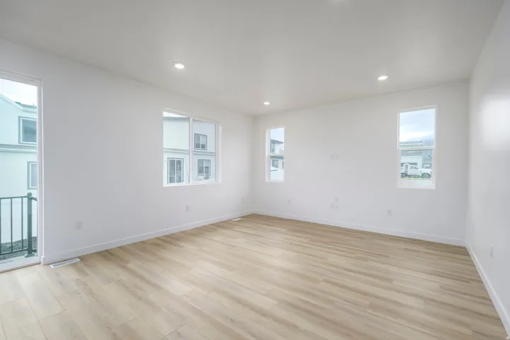 Spacious room featuring light wood-finish flooring, recessed lighting, multiple windows providing natural illumination, a sliding glass door with balcony access, and crisp white walls