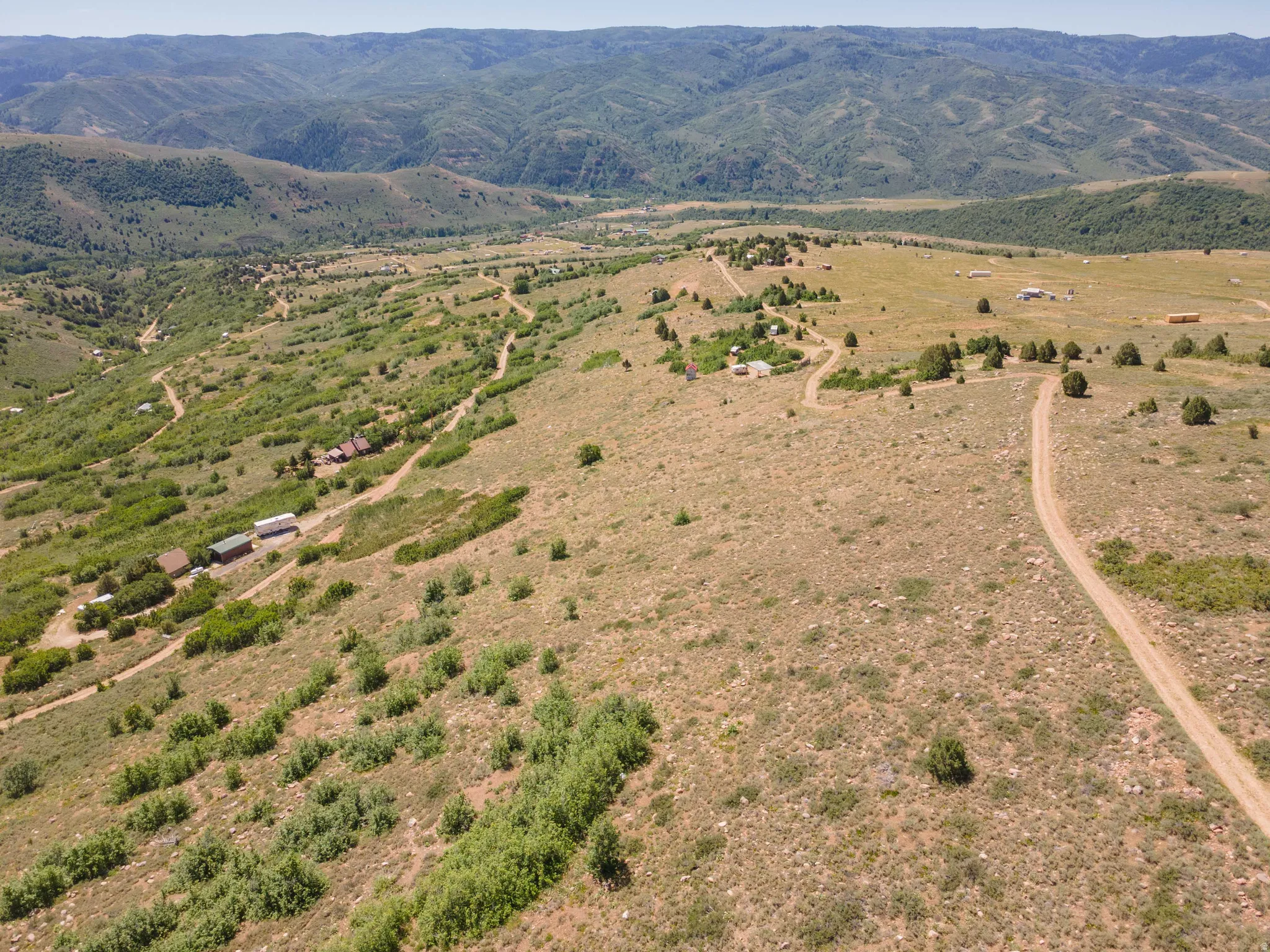 Expansive mountainous terrain featuring multiple structures, dirt roads, scattered trees, and distant valleys