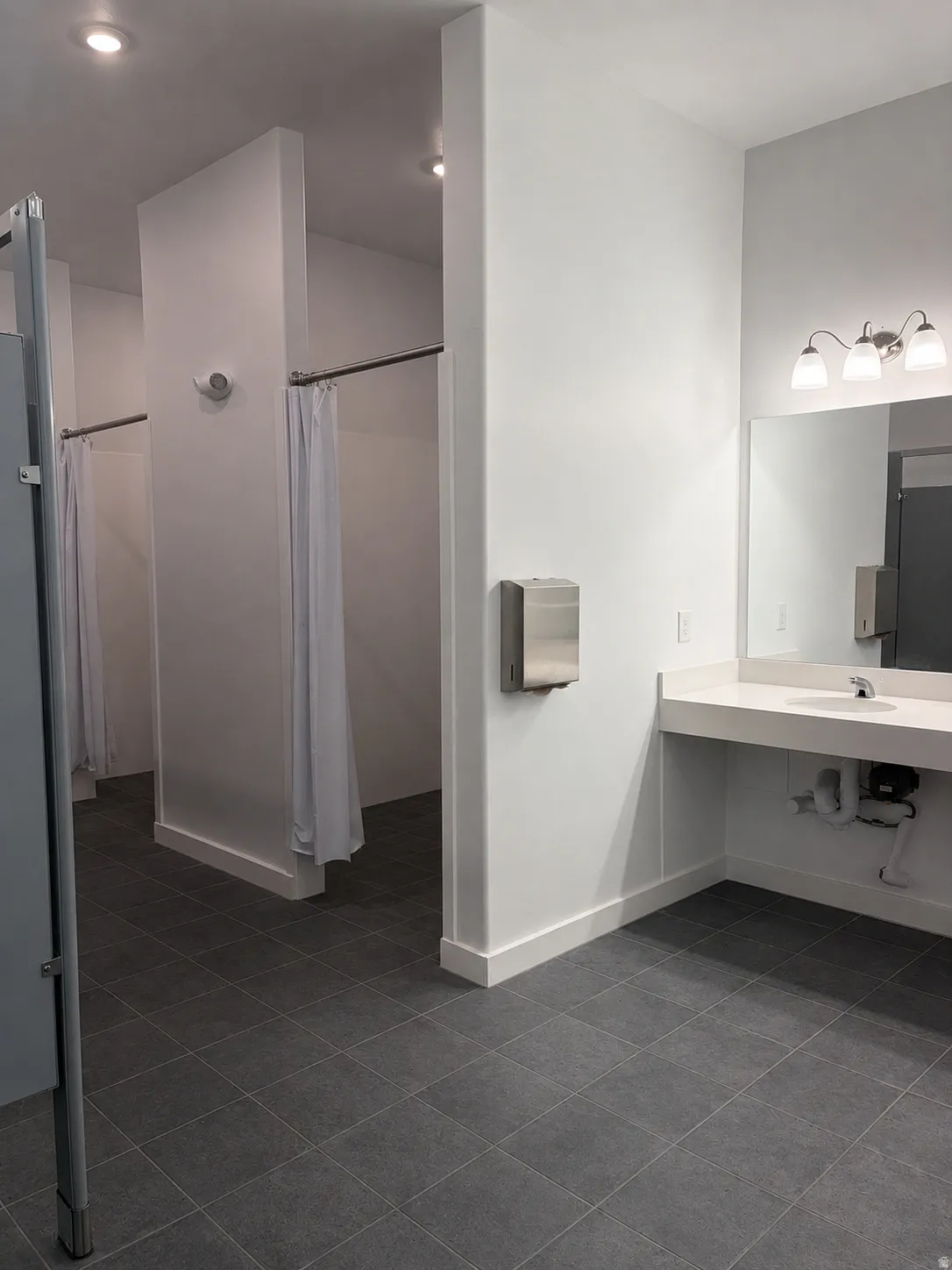 Commercial restroom featuring multiple shower stalls with privacy curtains, a vanity with an integrated sink, a large wall-mounted mirror, and tile flooring