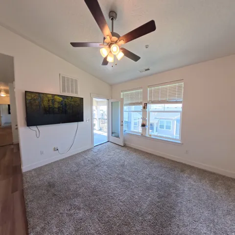 Carpeted interior space featuring a ceiling fan with integrated lighting, multiple windows with horizontal blinds, and a glass-paneled door opening to an exterior balcony