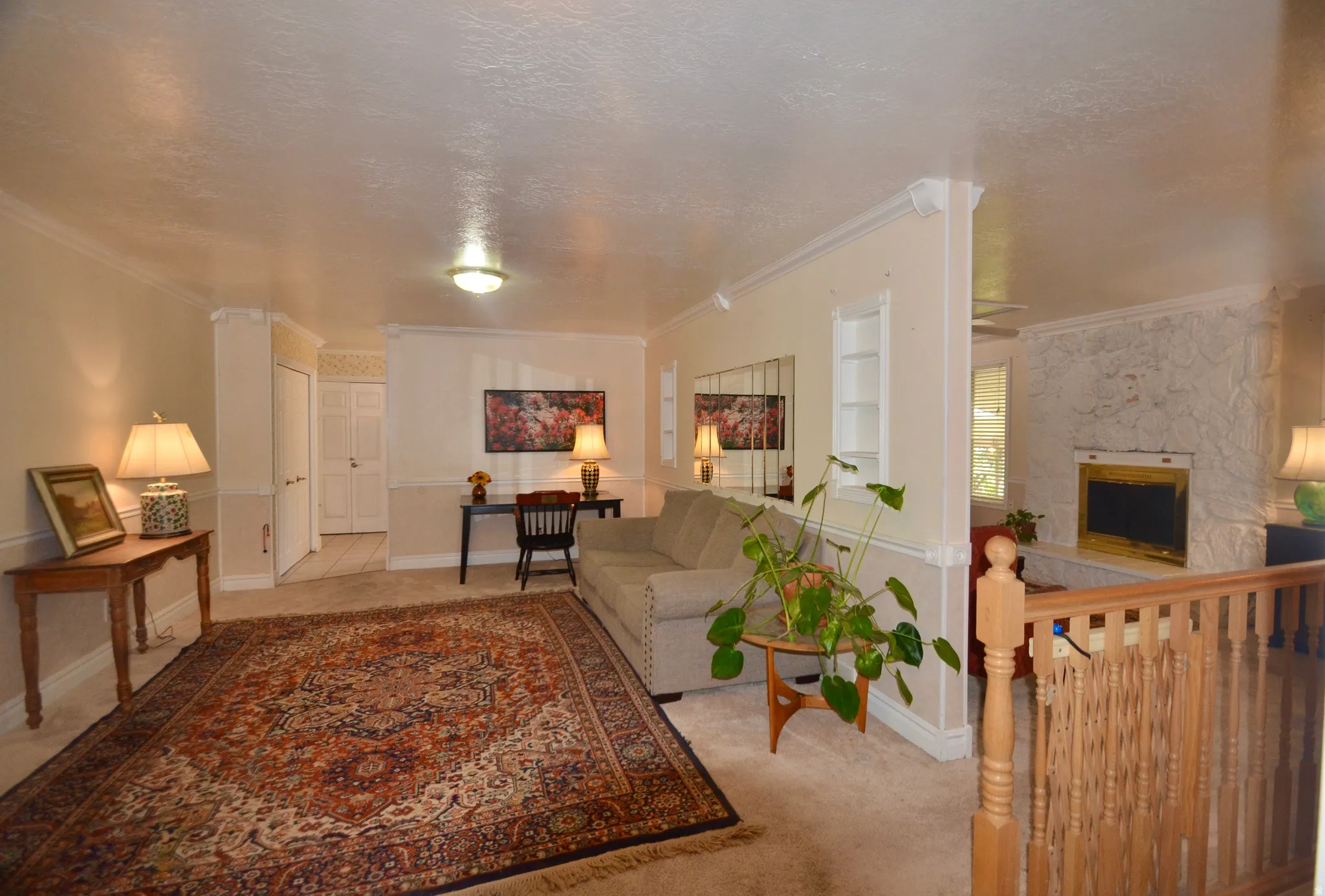 Carpeted living area featuring a decorative stone fireplace with a gold-tone insert, a wood banister, and crown molding