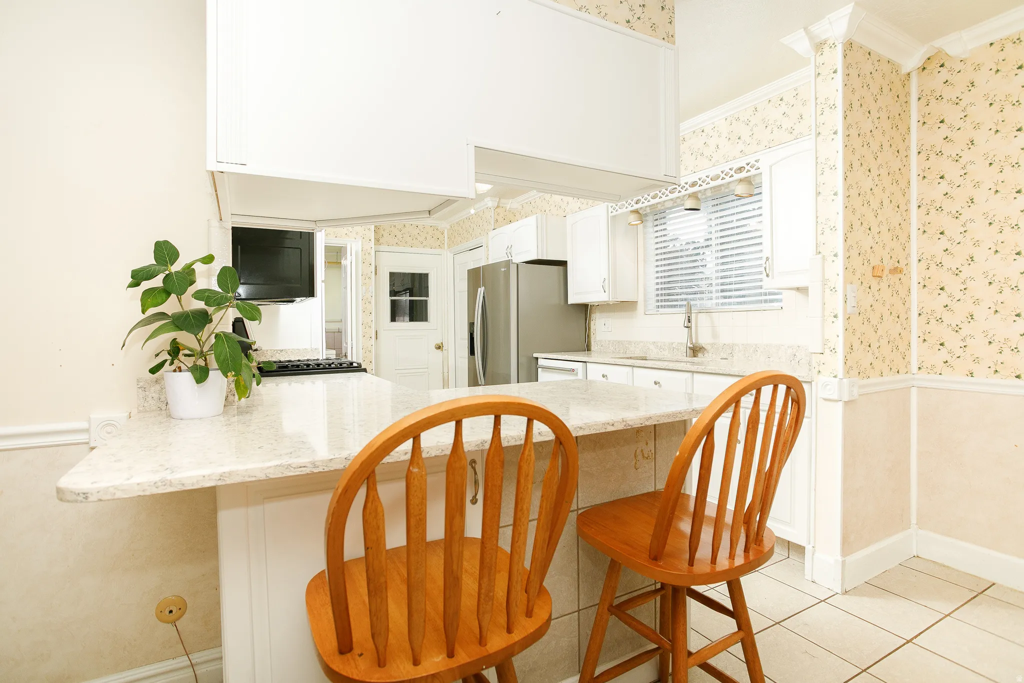 Kitchen featuring a peninsula breakfast bar with light-colored countertops, white cabinetry, a stainless steel refrigerator, tile flooring, and a window with horizontal blinds