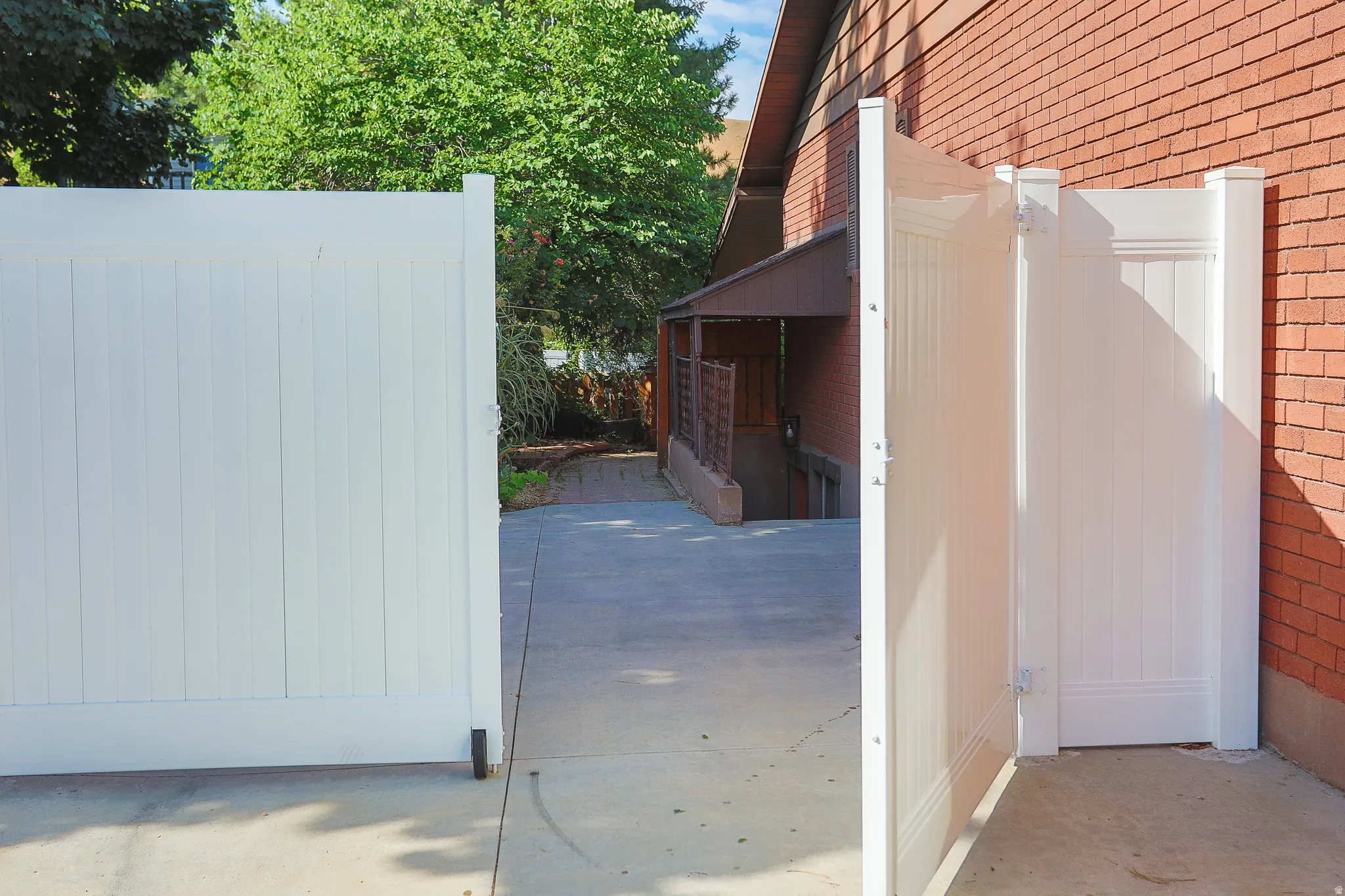 White vinyl privacy fence with an open gate, concrete pavement, and a red brick exterior