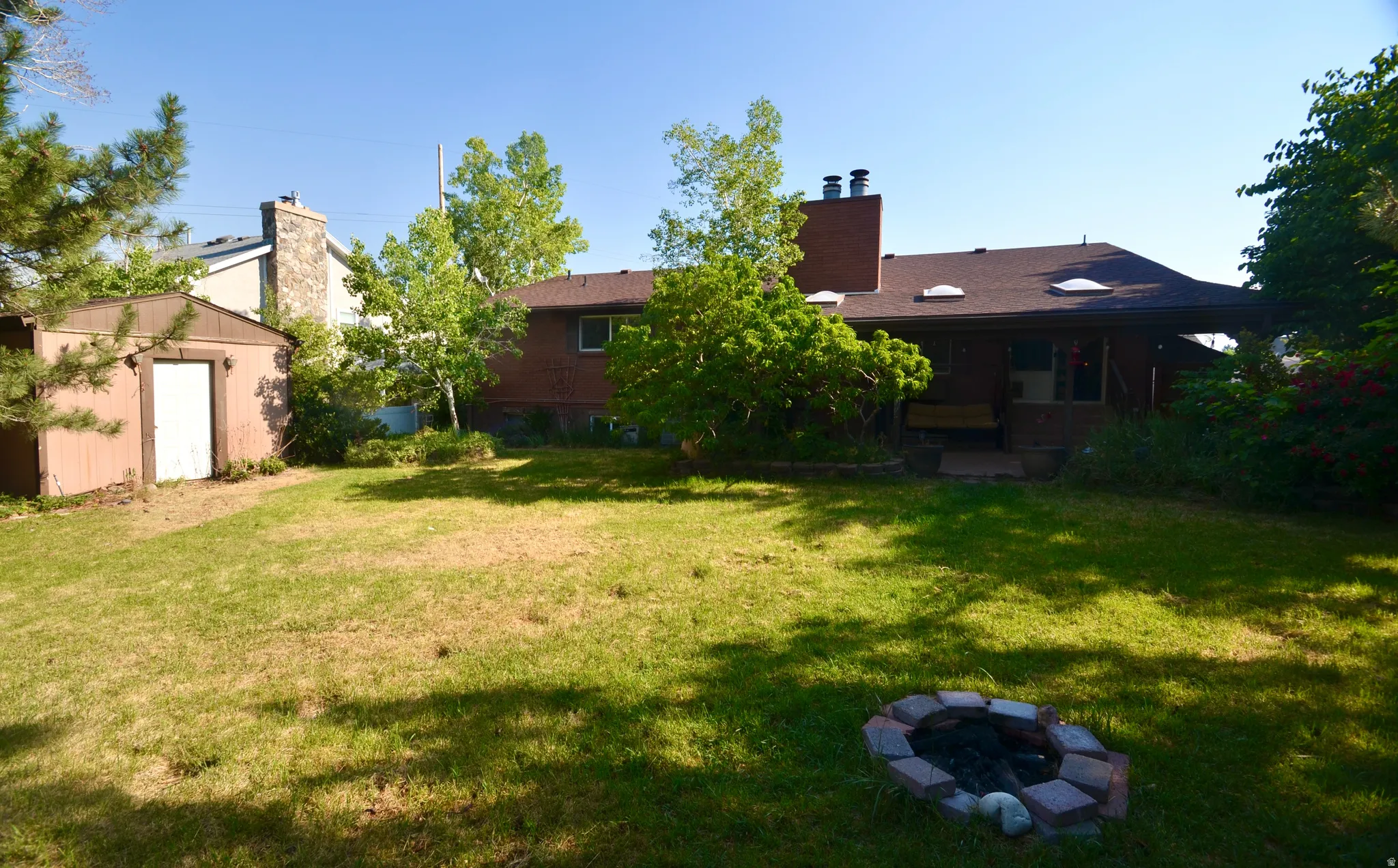 Expansive grassy backyard featuring a fire pit, mature trees, an outbuilding, and a covered patio area