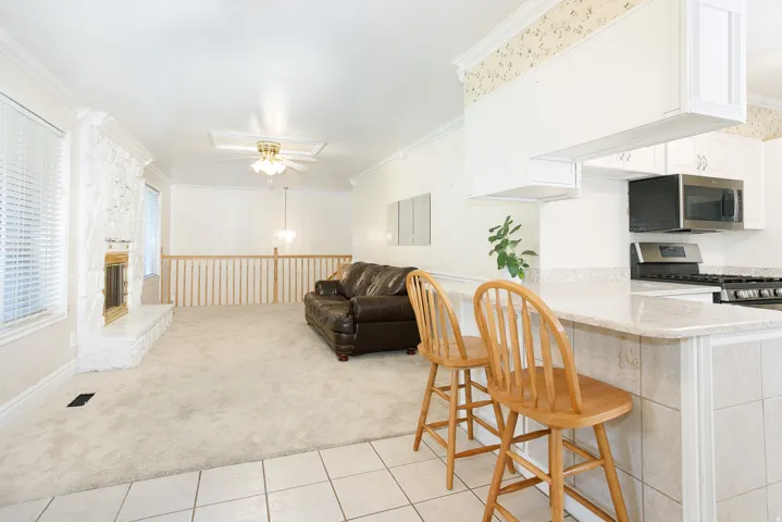Kitchen pass-through breakfast bar with tiled counter and base, opening to a carpeted living area featuring a stone-clad fireplace, ceiling fan, and spindle railing