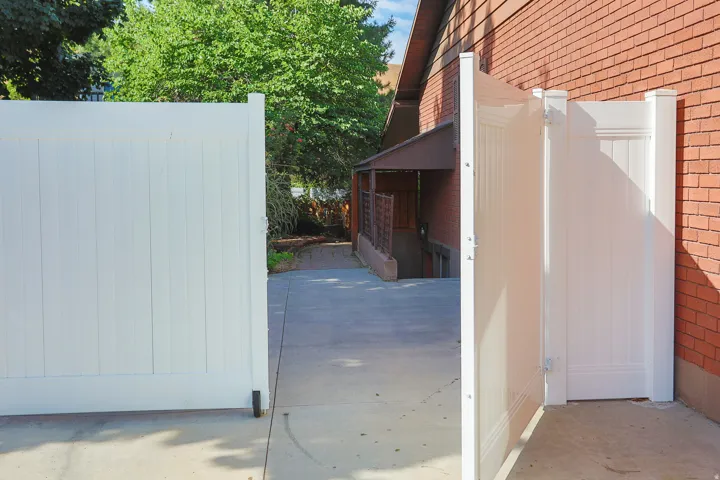 White vinyl privacy fence with an open gate, concrete pavement, and a red brick exterior