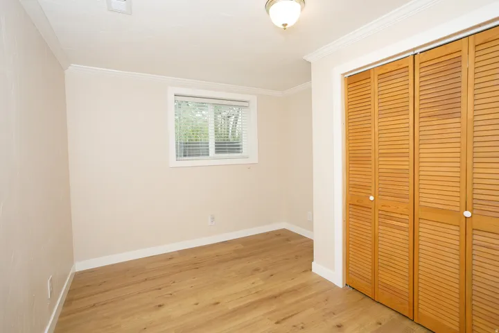 Room featuring wood-finish flooring, a window with blinds, bi-fold louvered doors, crown molding, and a ceiling-mounted light fixture