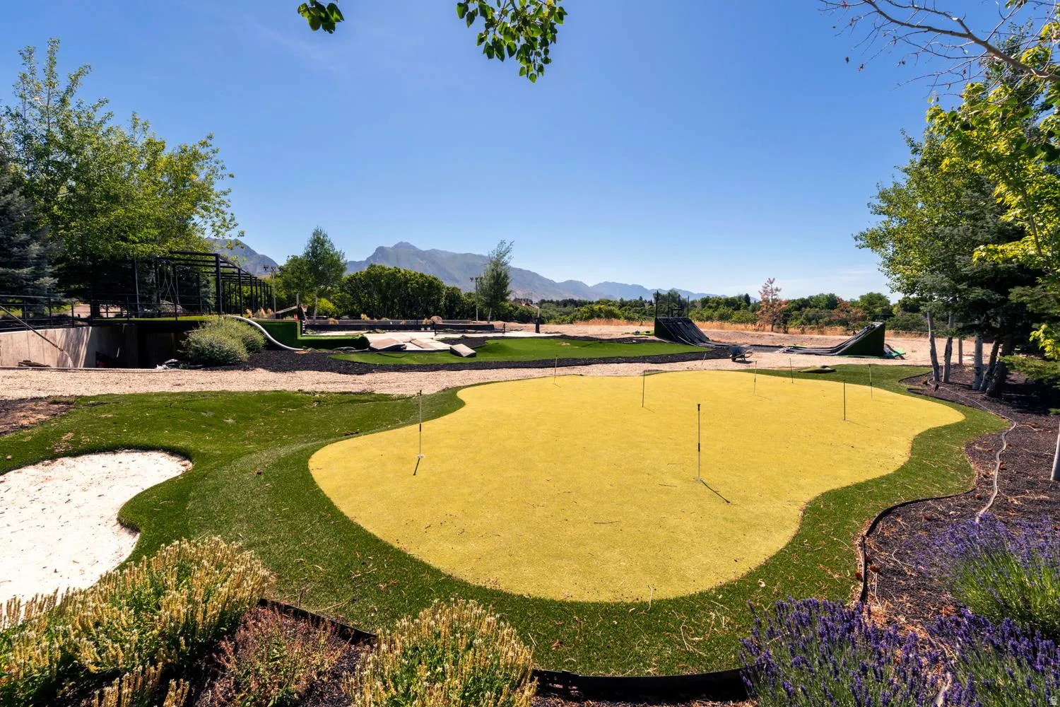 Putting green with multiple flag positions, bordered by a sand trap and landscaped shrubbery