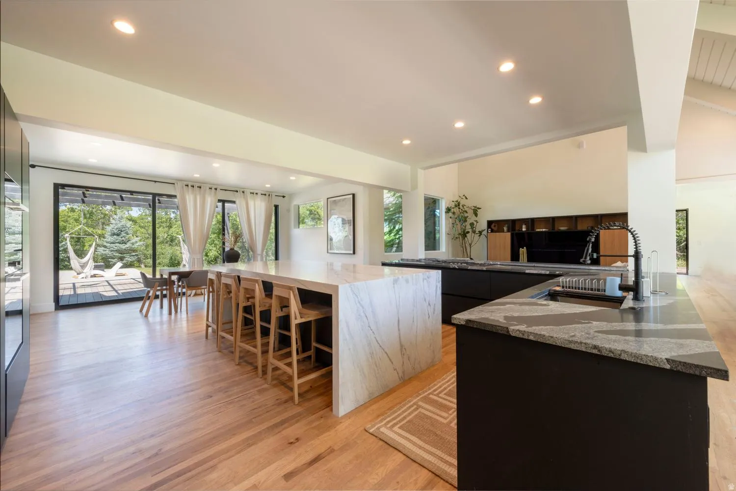 Open-concept kitchen featuring an oversized island with waterfall-edge countertop, wood-finish flooring, recessed lighting, black cabinetry, and a black pull-down faucet