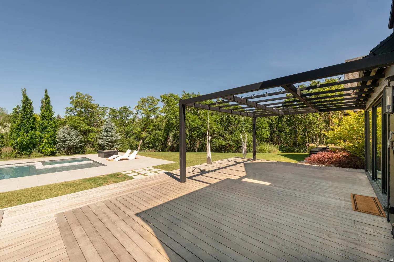 Expansive outdoor living area featuring a large wood-finish deck, a modern black pergola, and a rectangular swimming pool with integrated tanning ledge