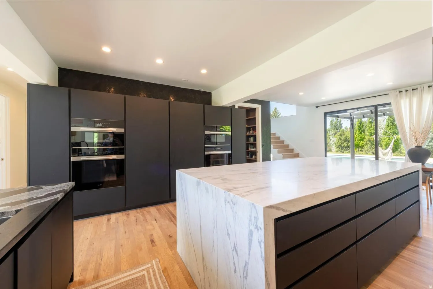 Sleek kitchen featuring a waterfall island with a marble countertop, integrated dark cabinetry, built-in ovens, and wood-finish flooring