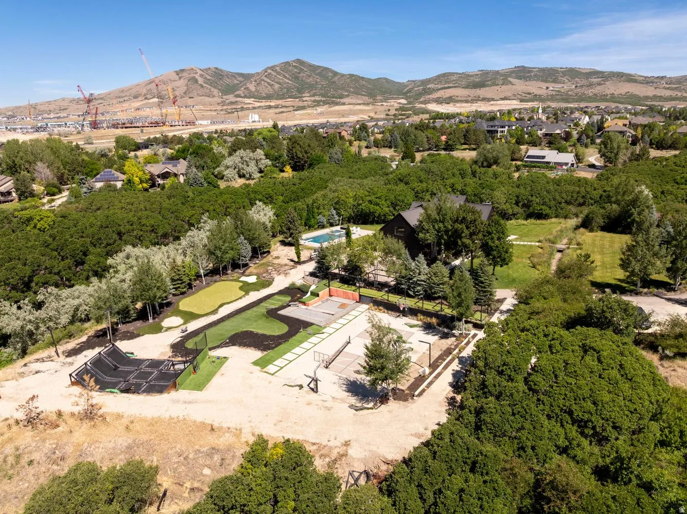 Expansive property featuring a dark-toned residence surrounded by mature trees, a swimming pool, a putting green, and a multi-sport court, all against a mountainous backdrop
