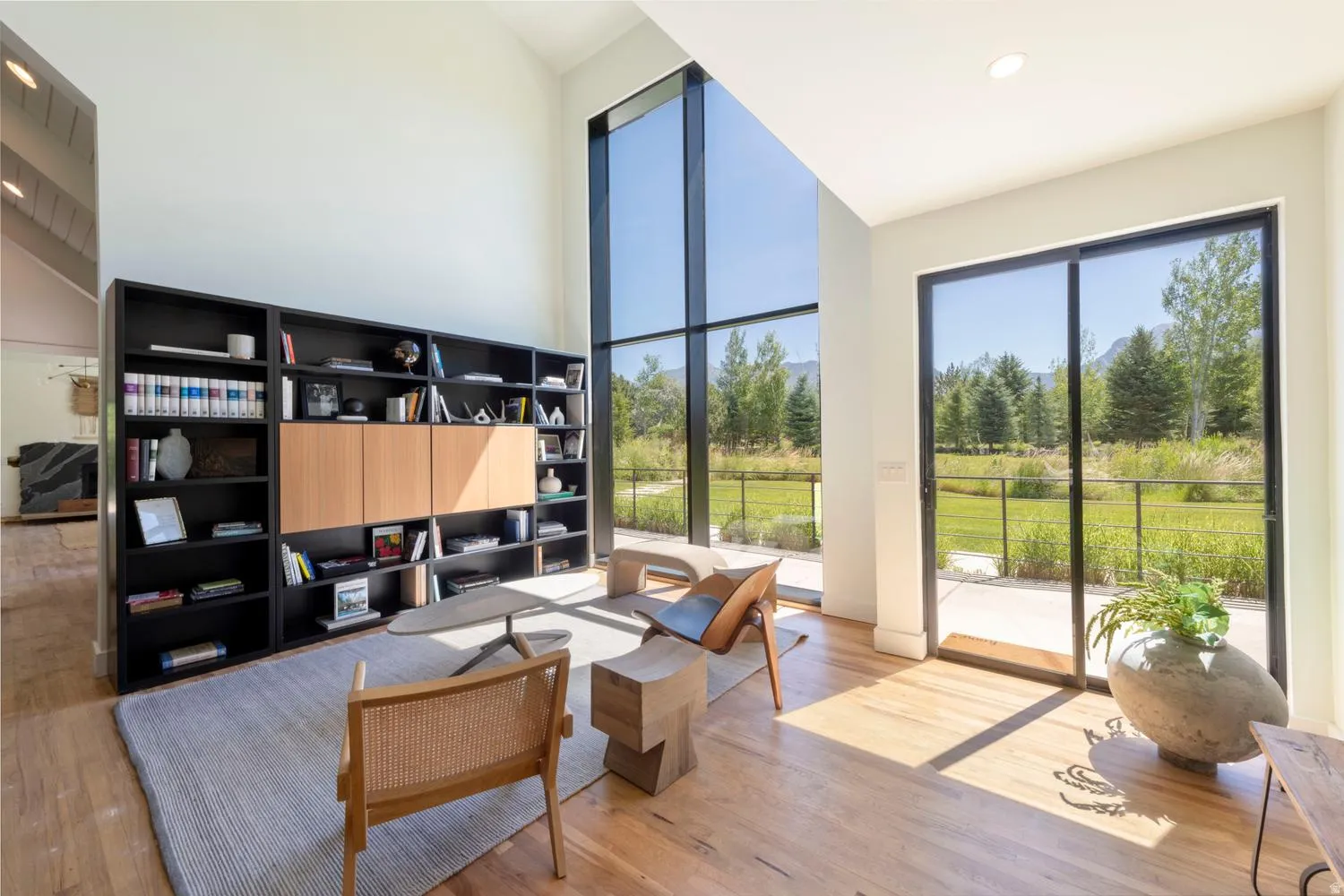 Spacious interior featuring a soaring ceiling, an expansive window wall, light wood-finish flooring, a built-in shelving unit with light wood cabinetry, and a sliding glass door opening to a patio
