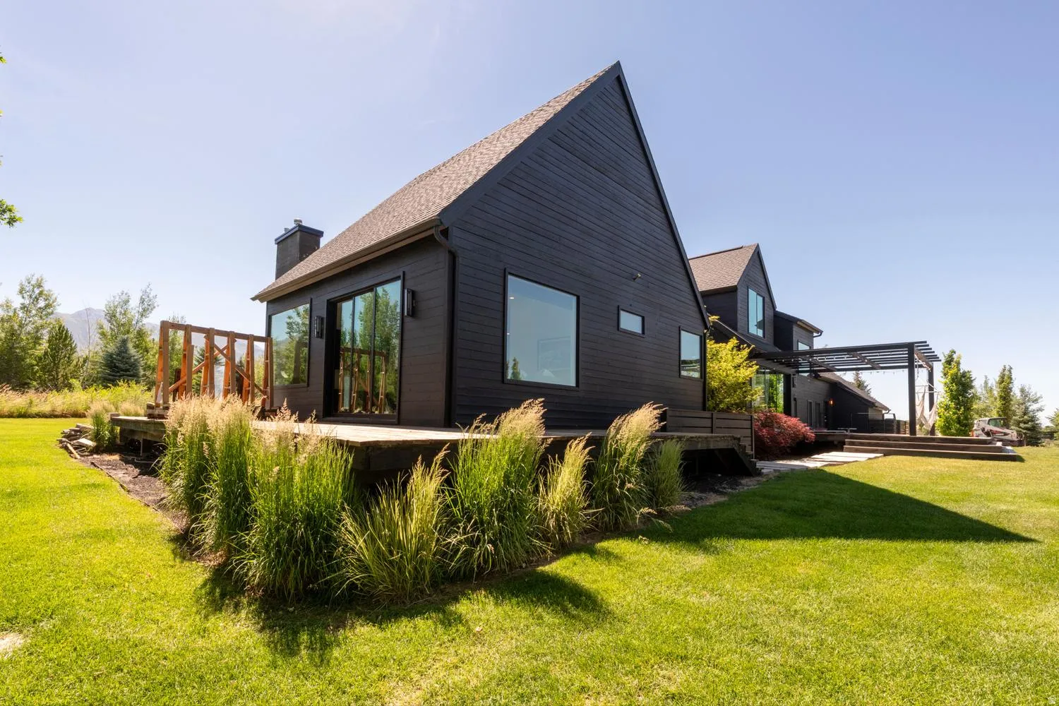 Modern black exterior featuring a prominent gable roofline, large window expanses, a spacious wooden deck with a partial railing, an integrated chimney, and a meticulously landscaped yard with mature plantings