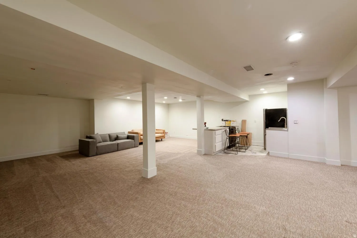 Open-concept finished lower level featuring recessed lighting, plush carpet flooring, a built-in wet bar with a sink, and a refrigerator recess