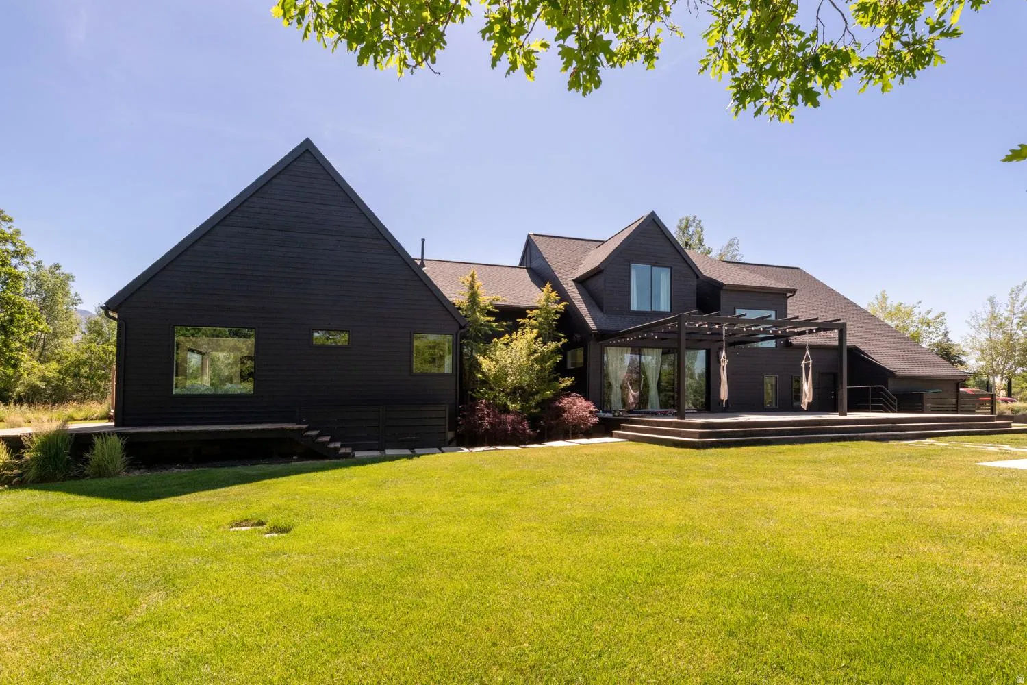 Expansive rear exterior featuring dark siding, deck, integrated pergola, large windows, and a generous lawn