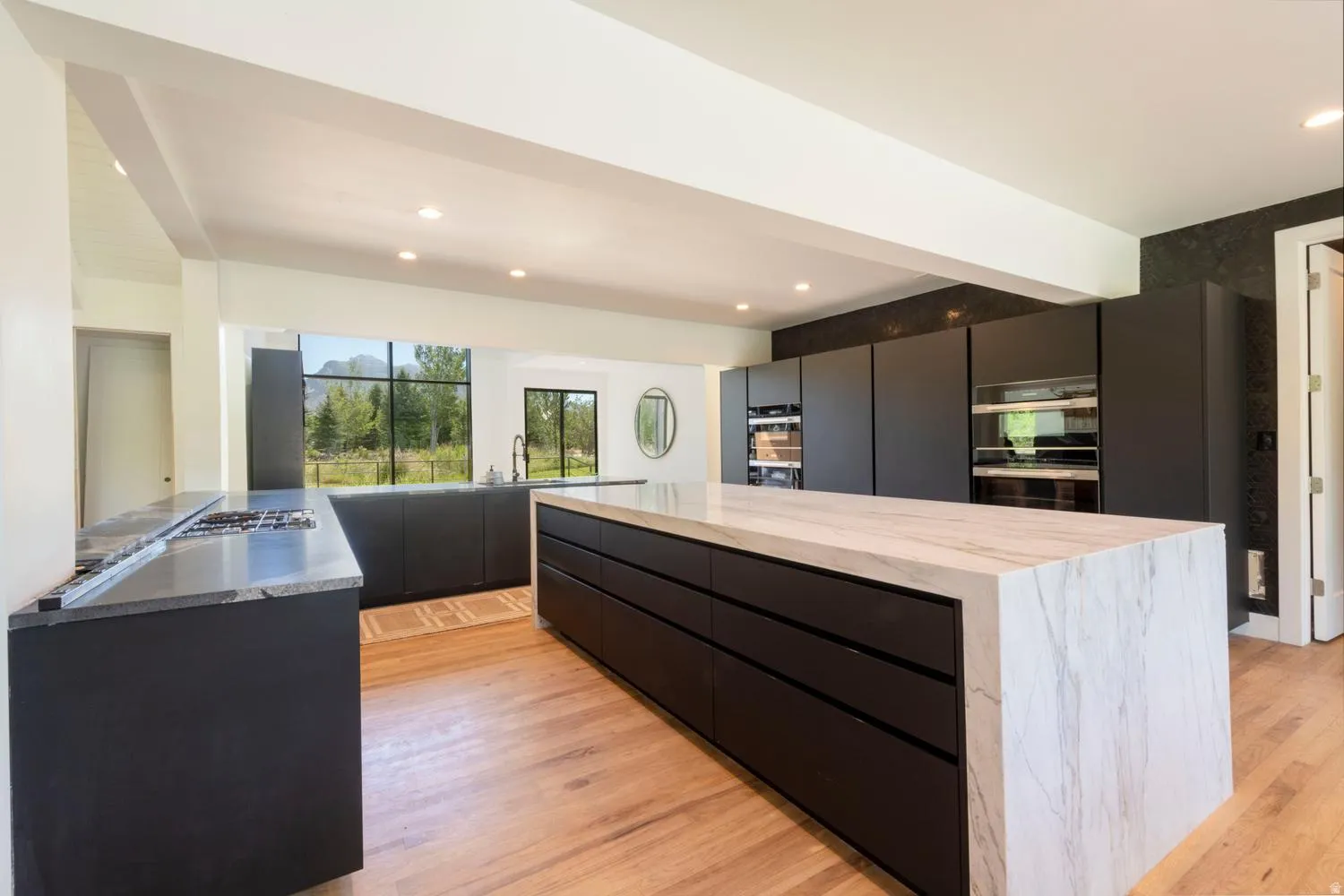 Contemporary kitchen featuring a large waterfall island with marble countertop, integrated dark cabinetry, multiple built-in ovens, a gas cooktop, and light wood-finish flooring