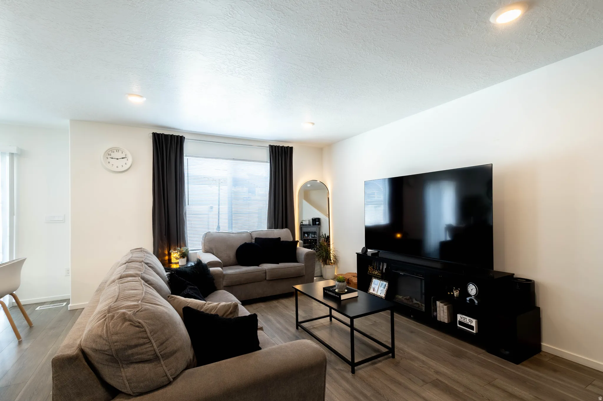 Spacious living area featuring light wood-finish flooring, recessed lighting, and a large window with privacy blinds and drapes