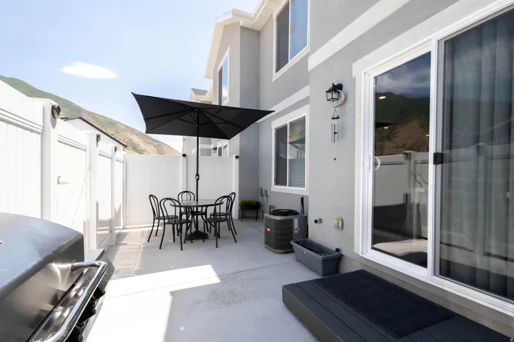 Paved patio area featuring privacy fencing, a gray stucco exterior, white-framed windows, a sliding glass door, and an exterior light fixture