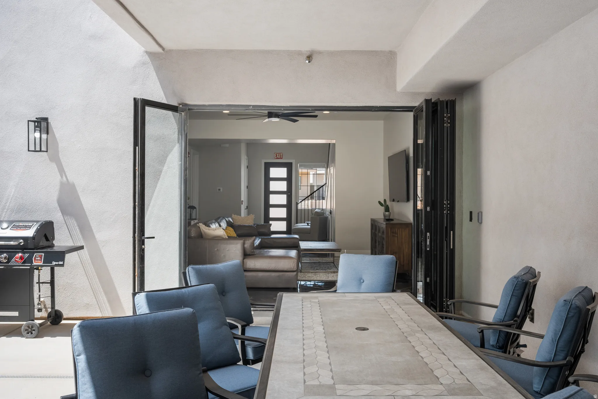 Exterior patio dining area featuring a tiled table surface, direct access to the interior via large folding glass doors, and a contemporary wall-mounted light fixture