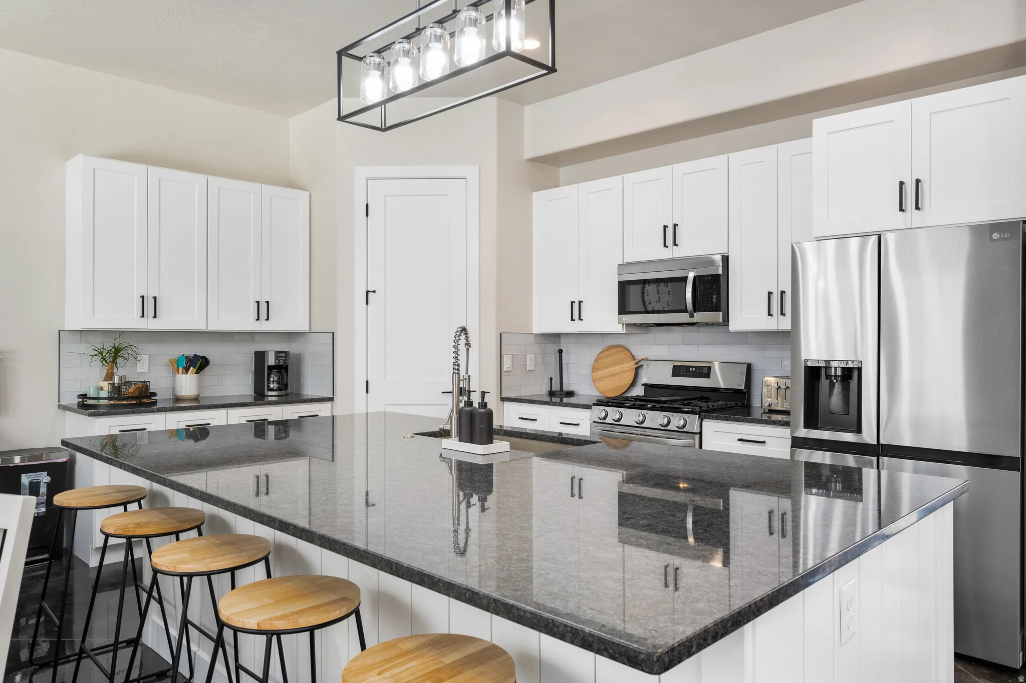Spacious kitchen featuring a large island with a dark countertop, white shaker-style cabinetry, stainless steel appliances, a tile backsplash, and a modern linear chandelier
